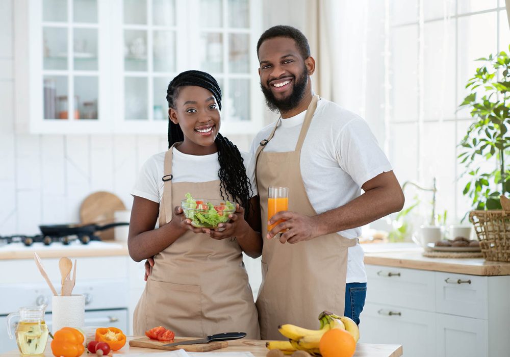 Vegan Family. Happy Black Couple With Fresh Vegetable Salad And Orange Juice Posing In Kitchen Interior, Young African American Spouses Enjoying Healthy Lifestyle And Tasty Food, Free Space